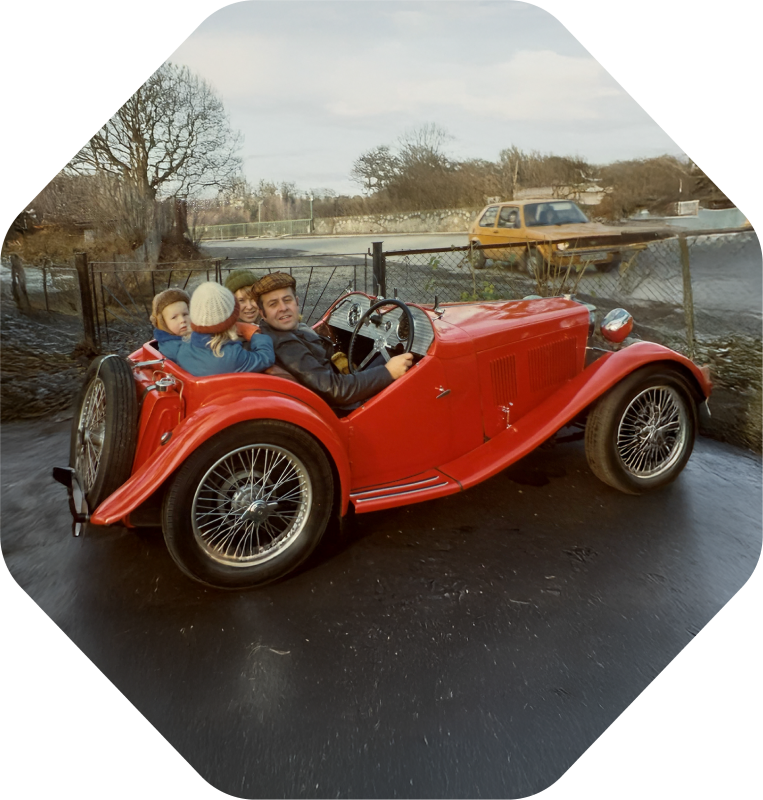 Old photo from the 70's showing Per Gjerdrum with his wife and kids all sitting in a parked vintage red MG T-type. A yellow VW Golf passes by on the road on the other side of a wire-fence.