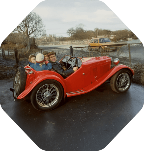 Old photo from the 70's showing Per Gjerdrum with his wife and kids all sitting in a parked vintage red MG T-type. A yellow VW Golf passes by on the road on the other side of a wire-fence. Old photo from the 70's showing Per Gjerdrum with his wife and kids all sitting in a parked vintage red MG T-type. A yellow VW Golf passes by on the road on the other side of a wire-fence.